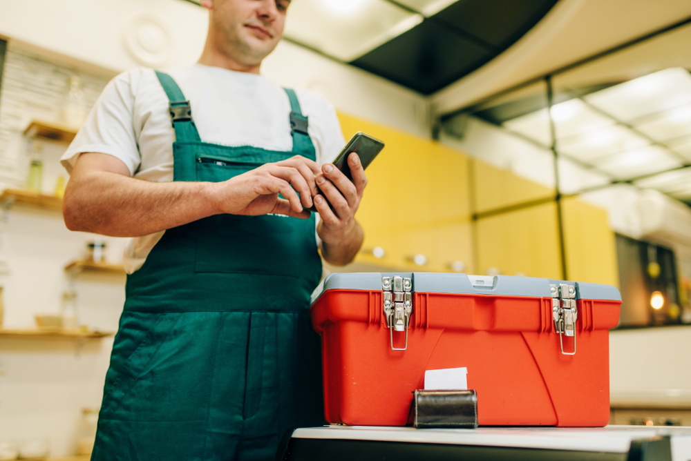 Repairman in uniform holding a phone next to a toolbox, representing the use of plumbing software for business management.